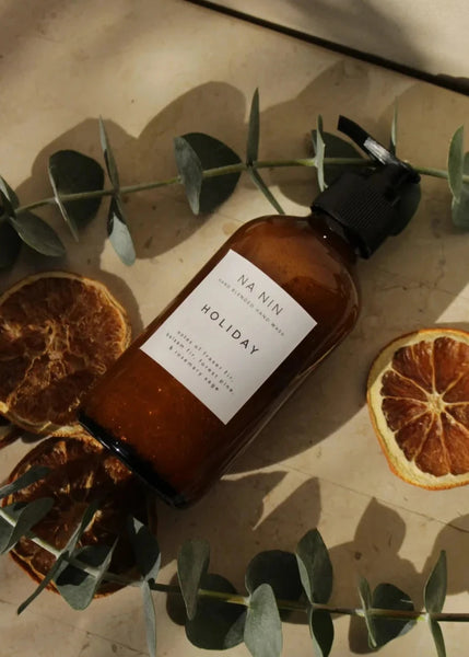 An 8oz amber glass bottle of hand wash with a pump dispenser, labeled 'Holiday', positioned on a surface with orange slices and green foliage in the background.