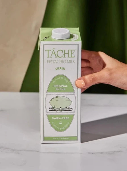 A hand with peach nail polish on places a carton of Táche pistachio milk on a marble table.