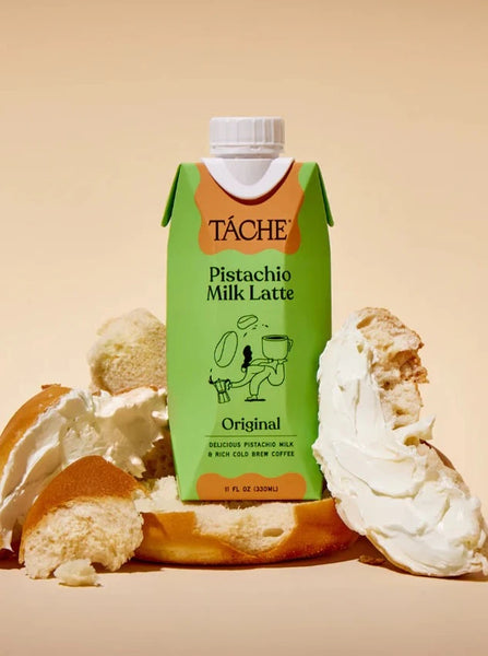 A carton of Táche pistachio milk latte sits on top of a crumbled bagel spread with cream cheese. 