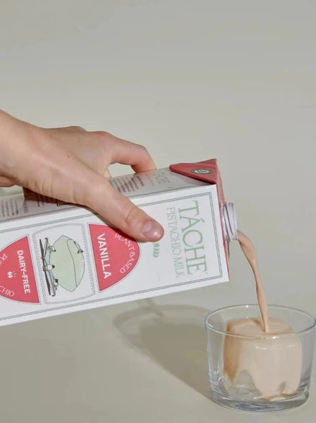 A hand pours a carton of Táche vanilla pistachio milk into a small glass with a large ice cube in it. 