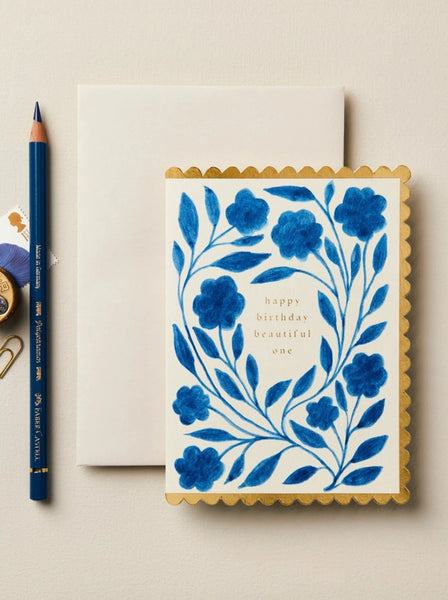 a blue floral card with a scalloped edge that says "happy birthday beautiful one" on a cream envelope next to a pencil and other desk accessories.