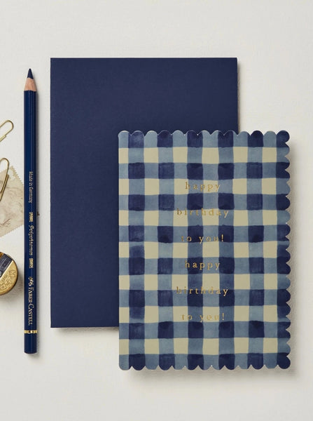 a blue and cream gingham card with a scalloped edge that says "happy birthday to you!" on a blue envelope next to a pencil and other desk accessories.