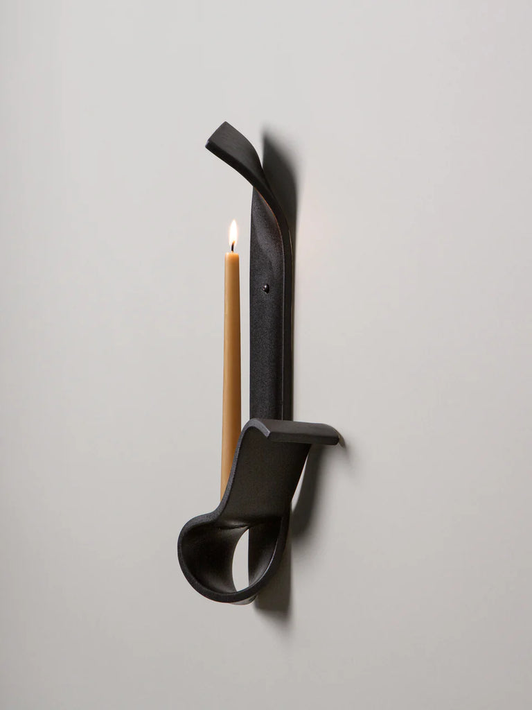 detail of a black ceramic wall sconce resembling a ribbon holds a lit beeswax taper on a white wall