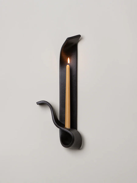 a black ceramic wall sconce resembling a ribbon holds a lit beeswax taper on a white wall