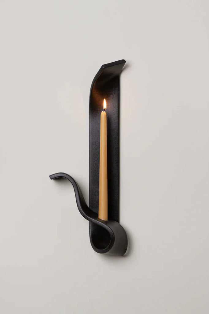 a black ceramic wall sconce resembling a ribbon holds a lit beeswax taper on a white wall