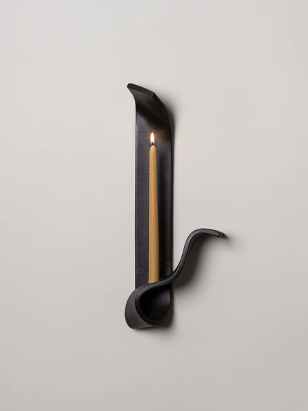 a black ceramic wall sconce resembling a ribbon made by sin ceramics holds a lit beeswax taper on a white wall