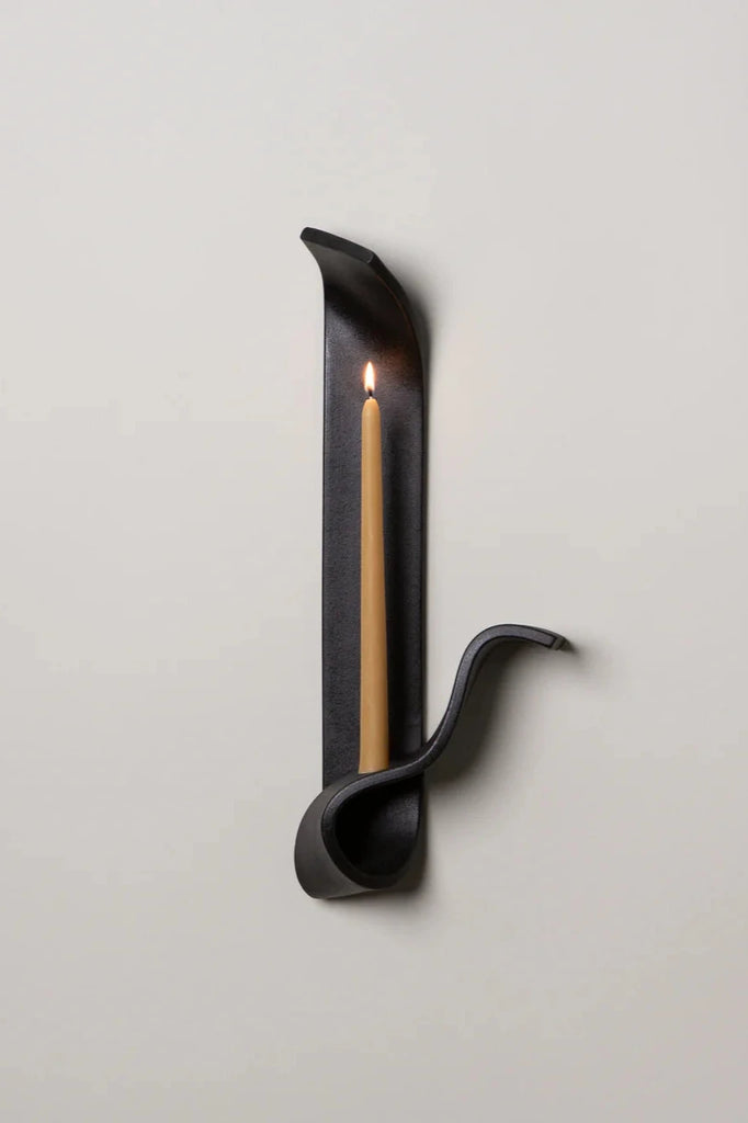 a black ceramic wall sconce resembling a ribbon made by sin ceramics holds a lit beeswax taper on a white wall