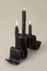 side view of black ceramic candelabra shaped like a ribbon with 3 lit candle tapers on a white background