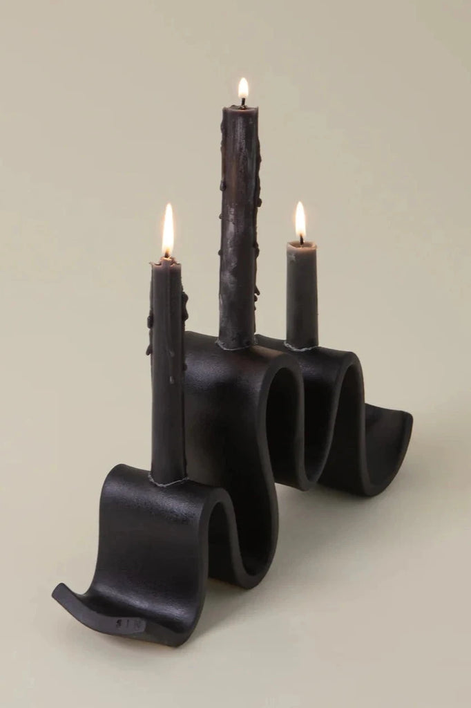 side view of black ceramic candelabra shaped like a ribbon with 3 lit candle tapers on a white background