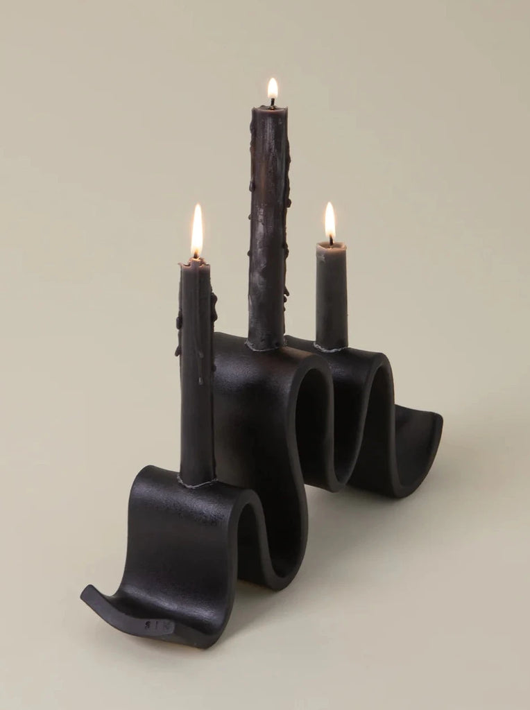 side view of black ceramic candelabra shaped like a ribbon with 3 lit candle tapers on a white background