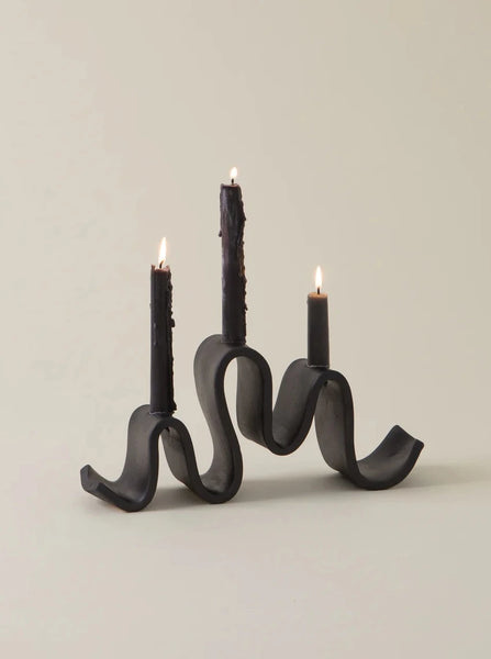 black ceramic candelabra shaped like a ribbon with 3 lit candle tapers on a white background