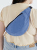 a woman wearing a pansy blue fanny pack with a black front zipper and black strap made by baggu over her shoulder and on her back against a white background