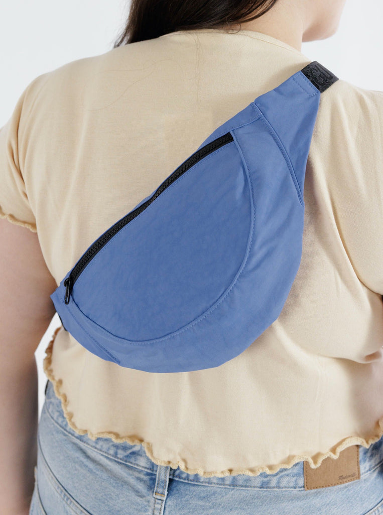a woman wearing a pansy blue fanny pack with a black front zipper and black strap made by baggu over her shoulder and on her back against a white background