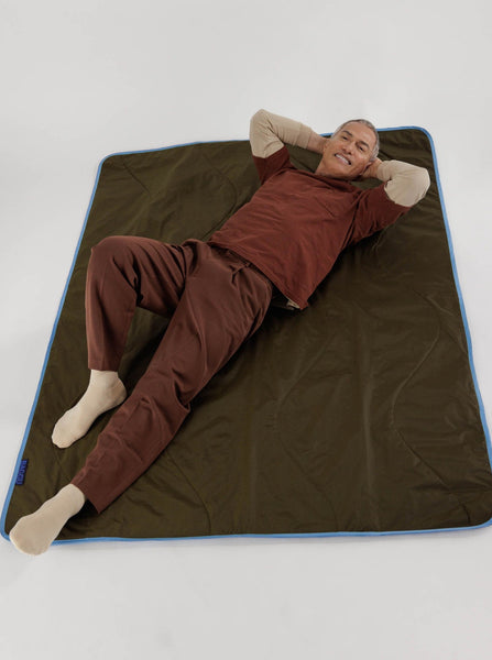 A guy laying on a baggu dark green puffy picnic blanket with baby blue trim against a white background
