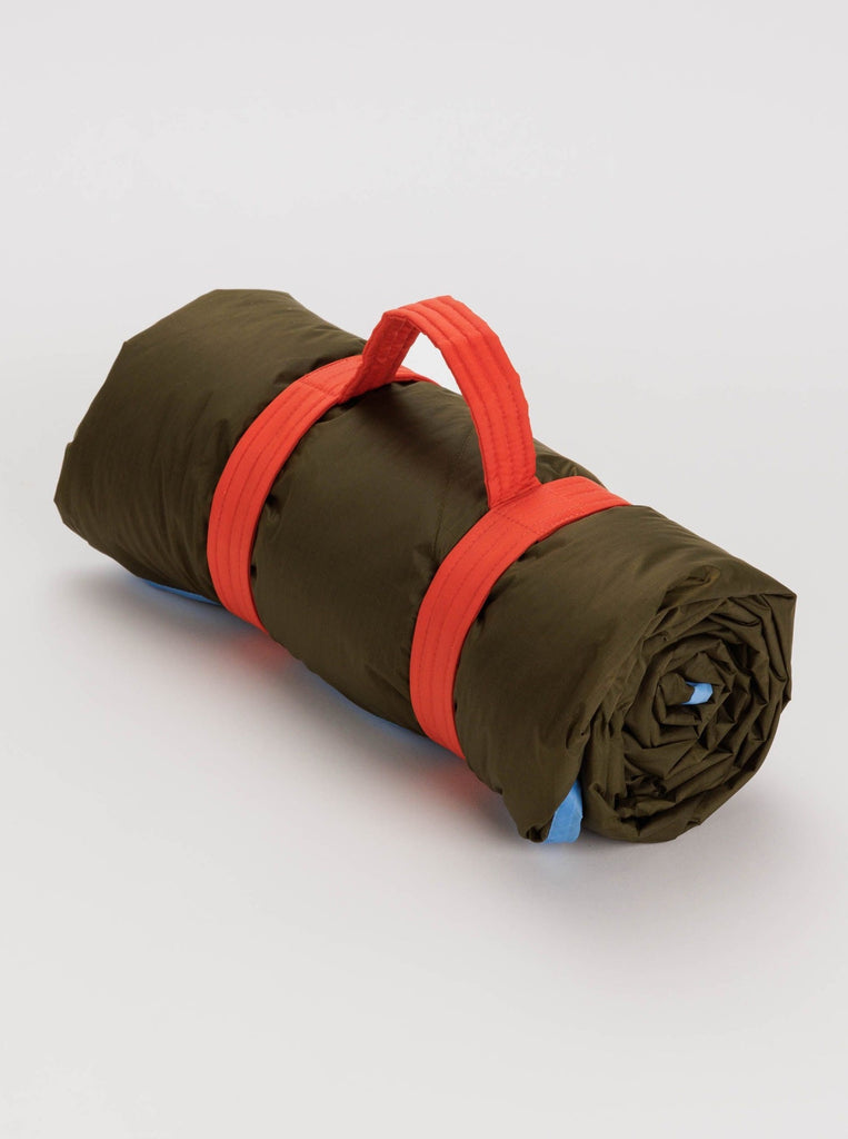 a dark green rolled up puffy picnic blanket with baby blue trim and an orange carrying handle made by baggu against a white background  Edit alt text