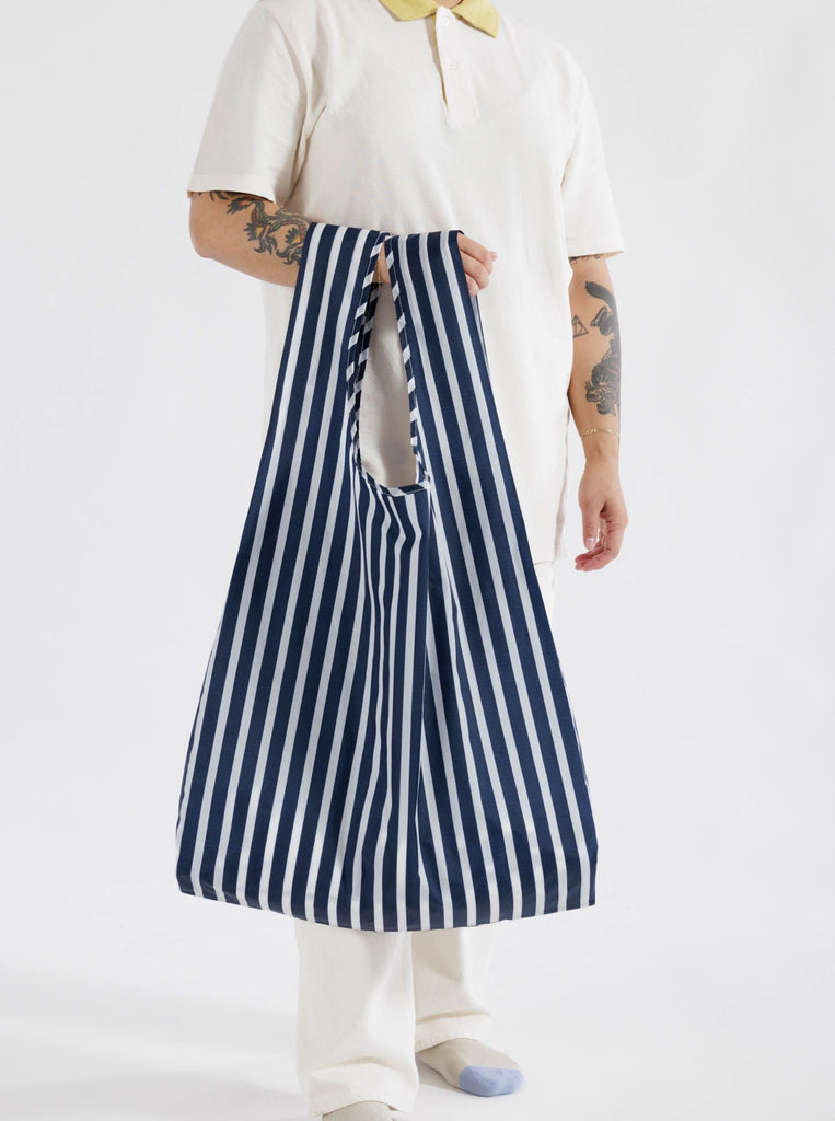 person with white shirt and tattoos carrying baggu reuseable big bag with navy and white stripes against a white background