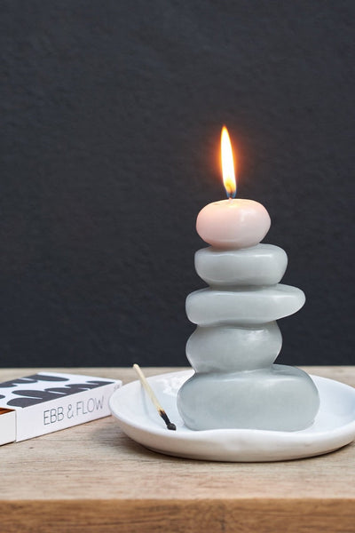 A lit light gray cairn shaped beeswax candle hand poured by claudia pearson sitting in a white dish beside an open box of ebb+flow matches against a dark background 