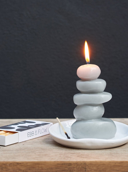 A lit light gray cairn shaped beeswax candle hand poured by claudia pearson sitting in a white dish beside an open box of ebb+flow matches against a dark background 