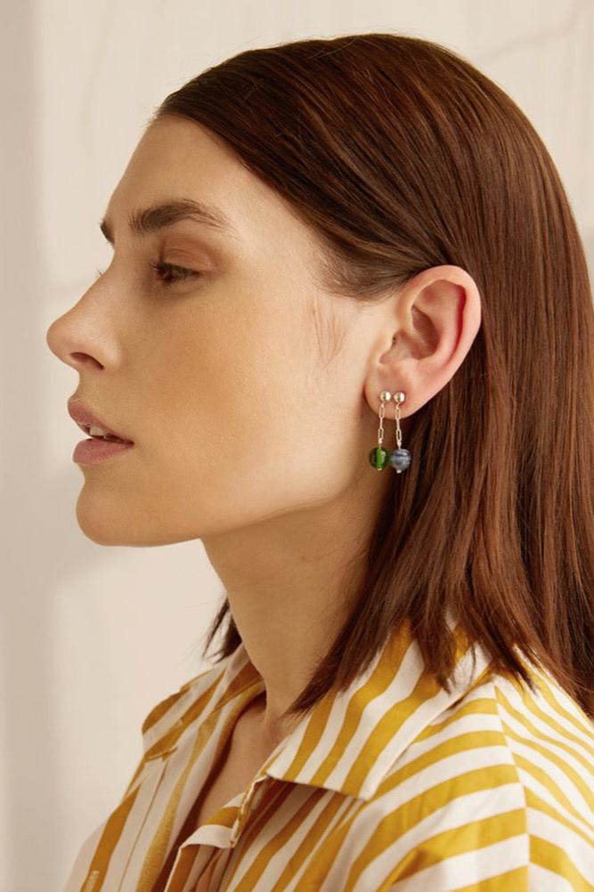 A woman wearing a yellow striped shirt and two earrings side by side each with a silver chain leading to a forest green gem and a dark and light blue swirl gem against a white background