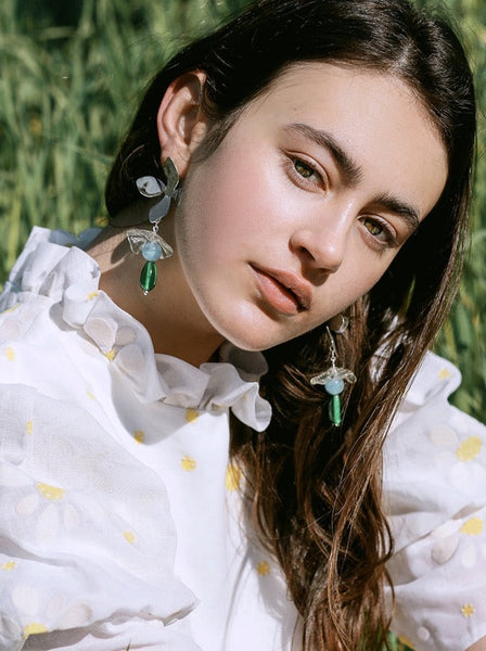 Woman in a white blouse sitting in grass wearing sterling silver flower shaped earrings with clear, baby blue and emerald green stacked eco gem glass beads made by cled Edit alt text