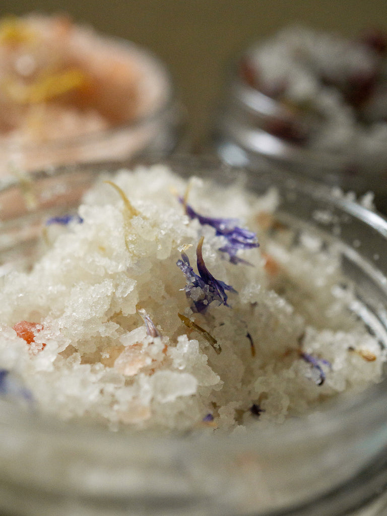 An open  jar of even keel bath salts with Organic Calendula petals,  Himalayan Pink and White Salts, Dead Sea salts, Sea salts, Magnesium Sulfate (Epsom), French Rose clay, Kaolin clay and pure Essential oils against a blurred background