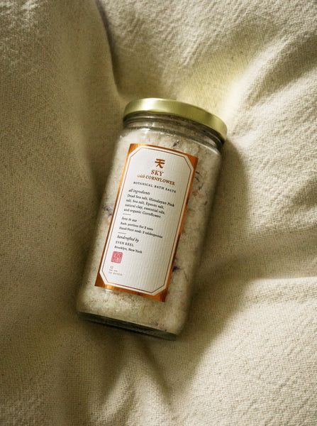 A glass jar of even keel bath salts against a beige fabric background