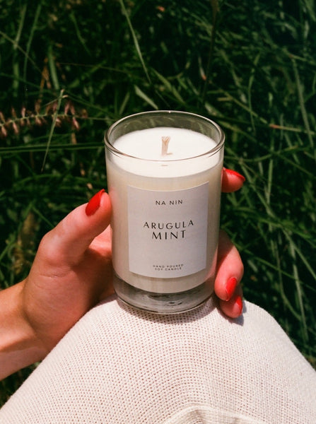 woman with red nails holding an arugula mint soy candle in glass container against a grass background