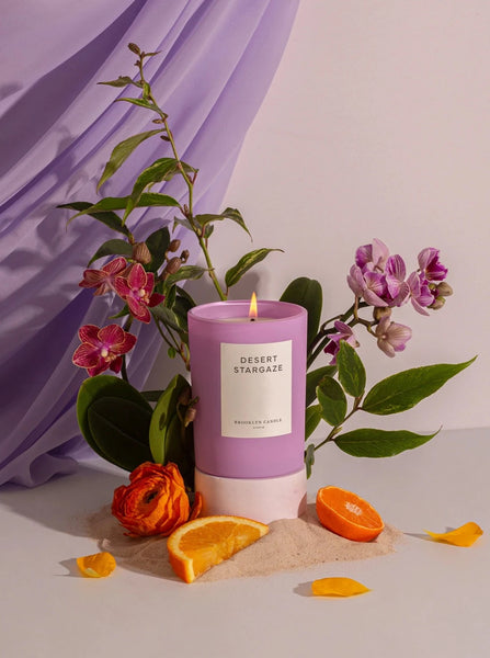 a frosted purple glass candle in the scent desert stargaze by Brooklyn candle company sits next to pieces of orange and orchid flowers